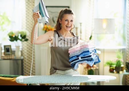 casalinga sorridente di 40 anni in blusa di seta e pantaloni beige con ferro a vapore, asse da stiro e un mucchio di vestiti stirati piegati nel moderno li Foto Stock