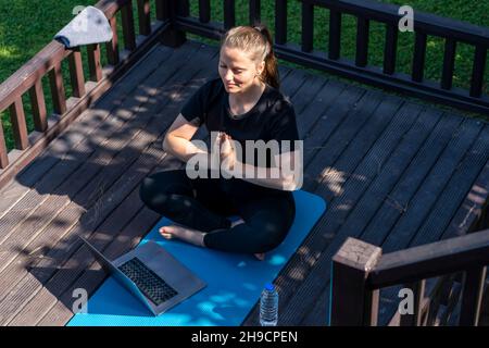 una donna sta facendo yoga sul portico della sua casa sorridendo meditazione nella posizione del loto. c'è un computer accanto ad esso classi online con un maestro Foto Stock