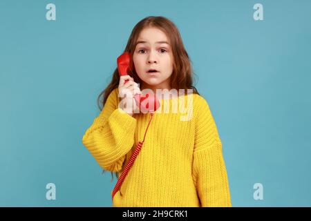 Ritratto di bambina tiene il telefono cellulare sorpreso dalla conversazione, scioccato da notizie incredibili, indossando giallo casual stile maglione. Studio interno girato isolato su sfondo blu. Foto Stock