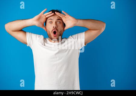 Ritratto di un giovane confuso che tiene le mani sulla testa su sfondo blu Foto Stock