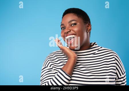 Donna africana urlando mano di tenuta vicino alla bocca giocosamente, sfondo blu Foto Stock