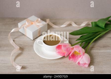 Caffè con latte in una tazza di porcellana bianca e piattino, un bouquet di tulipani rosa primaverile e un regalo in una scatola bianca con un nastro di raso. Festa della mamma, Val Foto Stock