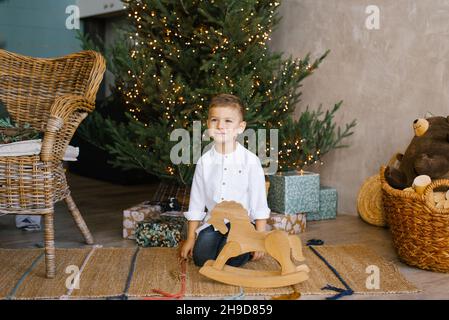 Il ragazzino carino è seduto vicino ad un albero di Natale e ad un cavallo a dondolo di legno, indossando una camicia bianca Foto Stock