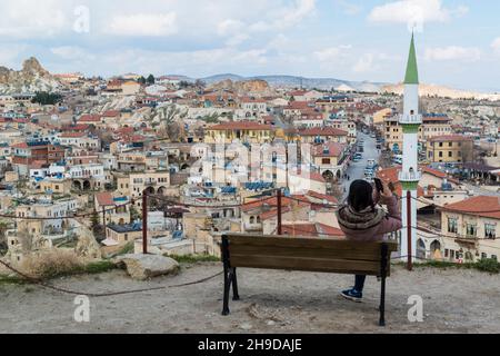 Turista femminile, seduto su una panchina, fotografando la città di Ortahisar con il suo smartphone da un punto di vista. Cappadocia, Turchia. Foto Stock