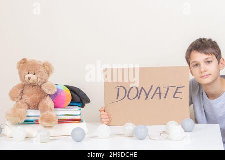 Beneficenza, donazione. Sorridente capretto che tiene pezzo marrone di cartone con iscrizione Donate. Volontario raccogliere vestiti, libri e giocattoli per la donazione. Foto Stock