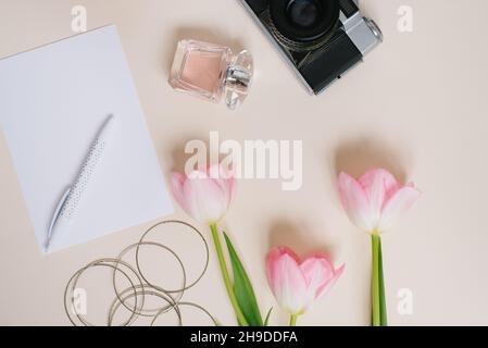 Macchina fotografica vintage, tulipani rosa primaverili, taccuini, un foglio bianco di carta e una penna, profumo e bracciali da donna su sfondo beige. Il concetto di s Foto Stock