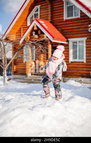 Il bambino è in piedi al cottage di legno. Bambina in inverno vestito caldo gioca in aria fresca e gode giorno di sole in inverno, lei si trova nella neve e giochi Foto Stock