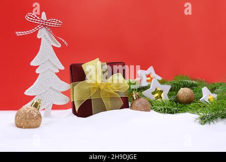 Albero di Natale con scintille, scatola di borgogna con arco giallo, palle d'oro lucido, lampadine a stella, rami di abete sulla neve su sfondo rosso. Anno nuovo. Foto Stock