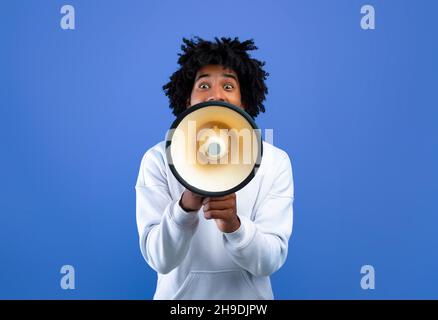 Emotivo teenager nero che fa annuncio con megaphone su sfondo blu studio Foto Stock