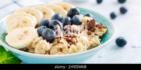 Porridge di avena con banana, cioccolato e mirtillo fresco in una ciotola su un fondo di ardesia, pietra o cemento grigio chiaro. Vista dall'alto. Foto Stock