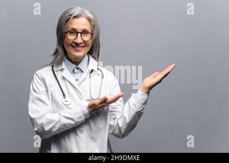 Un affascinante medico pediatrico femminile, medico, terapeuta che indossa un abito medico bianco con uno stetoscopio sulle spalle punta le mani su spazio vuoto, mock up, spazio copia. Sanità e medicina Foto Stock