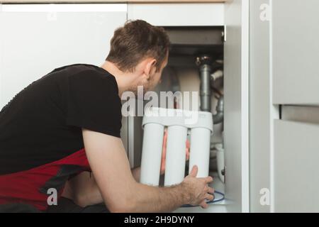 Idraulico in cucina installa un sistema di trattamento delle acque domestiche. Sistema per fiasche a tre stadi Foto Stock