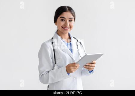 Allegro fiducioso millenario medico signora indiana in cappotto con tablet guardare la fotocamera, isolato su sfondo bianco Foto Stock