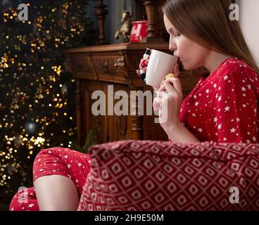 Giovane donna con tazza di tè calda seduta in sedia a casa accogliente durante le vacanze invernali. Persona in pigiama rossa in atmosfera moderna di Capodanno. Foto Stock