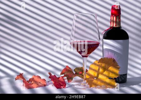 Bicchiere da vino e bottiglia con vino rosso e foglie d'uva autunnali su sfondo bianco con ombre cieche. Concetto romantico con ombre jalousie. Foto Stock