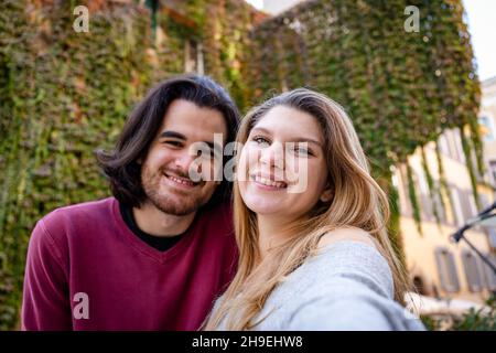 Bella coppia che viaggia a Roma. Una giovane coppia prende un selfie in una strada nel centro storico. Sullo sfondo un edificio coperto di edera. Foto Stock