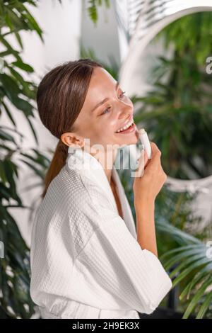 Ritratto di giovane donna carina con bella pelle sorridente mentre si applica il balsamo labbro sulle labbra, in piedi in bagno Foto Stock