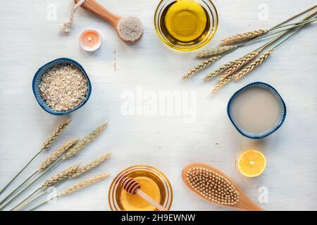 Ingredienti naturali per scrub fatto in casa. Fiocchi di avena, olio, latte di avena, miele, limone, spazzola per viso e corpo e orecchie di grano su tavola di legno bianco. Vista dall'alto Foto Stock