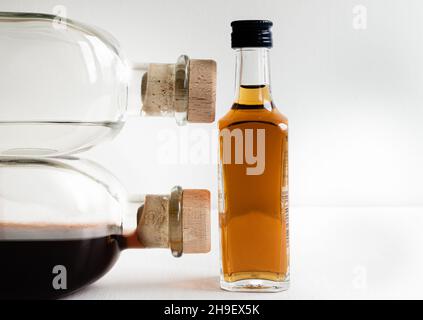 Due grandi bottiglie di vetro con liquidi e tappi contro una piccola con cognac su sfondo sfocato chiaro Foto Stock