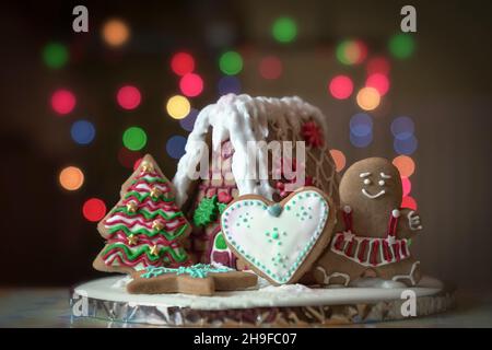 Casa di pan di zenzero decorato in casa, albero, donna e biscotti a forma di cuore sul tavolo con bokeh. Foto Stock