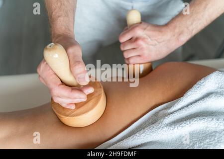 Primo piano sul retro della gamba di donna sconosciuta che ha madero terapia massaggio anti-cellulite trattamento da terapista professionista che tiene in mano strumenti in legno Foto Stock