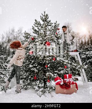 Giovane coppia felice in elegante abbigliamento invernale decorando albero di Natale con palle all'aperto nella foresta innevata d'inverno Foto Stock