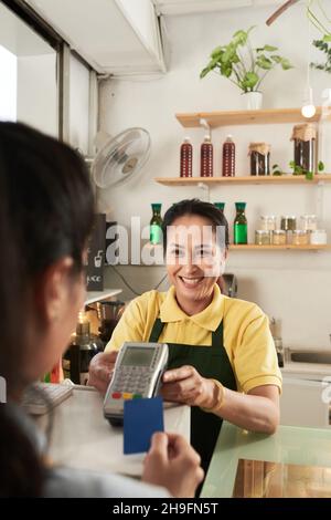 Il lavoratore allegro del caffè accetta il pagamento e offre il terminale di pagamento al cliente Foto Stock