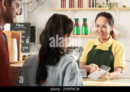 Barista femminile allegra matura che mostra il prezzo dell'ordine sul calcolatore ai clienti Foto Stock