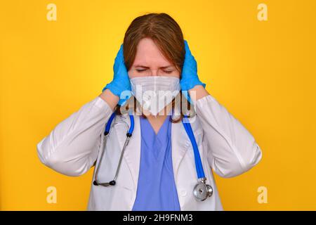Il medico tiene le mani sopra la testa su uno sfondo giallo, mal di testa. Infermiera in uniforme medica - stress e affaticamento dovuto coronavirus Foto Stock