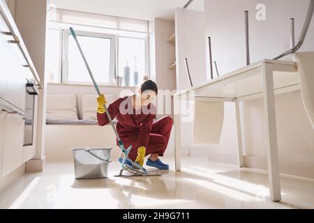 Janitor focalizzato in guanti di gomma seduti su fianchetti e montaggio mop in microfibra in cucina Foto Stock