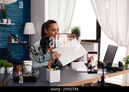 Donna presentatore filma vlog revisione del computer portatile. Influencer sui social media che fa consigli sui gadget di fronte alla fotocamera dello smartphone. Trasmissione di podcast online tramite tecnologia. Foto Stock