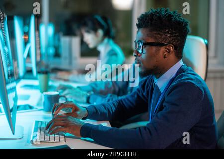 Giovane uomo d'affari serio in occhiali e formalwear guardando lo schermo del computer contro i suoi colleghi Foto Stock