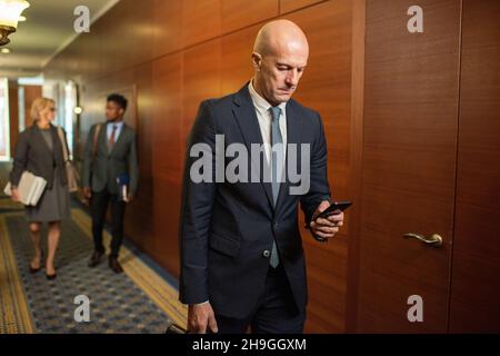 L'avvocato mature calvo scorre in smartphone mentre si sposta lungo il corridoio di un grande centro di uffici contemporaneo Foto Stock