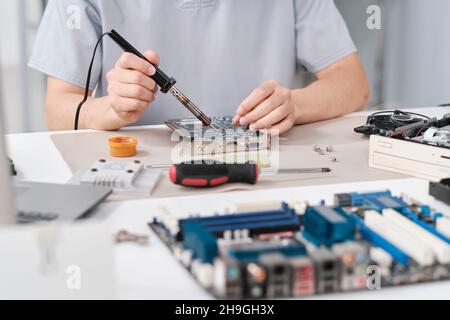 Mani di un giovane addetto alla riparazione che fissa piccoli dettagli sulla scheda madre con saldatore Foto Stock