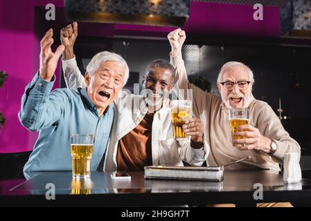 eccitati amici multietnici anziani che mostrano il gesto di trionfo mentre si guarda il campionato di calcio in pub Foto Stock