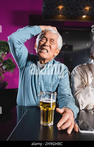 scioccato uomo asiatico senior con occhi chiusi che toccano la testa mentre si siede con un amico afroamericano nel bar dello sport Foto Stock