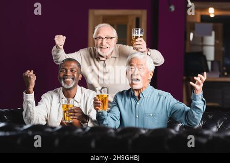 amici multietnici anziani allegri che tengono la birra e mostrano il gesto di vincere mentre guardano la partita di calcio al bar Foto Stock