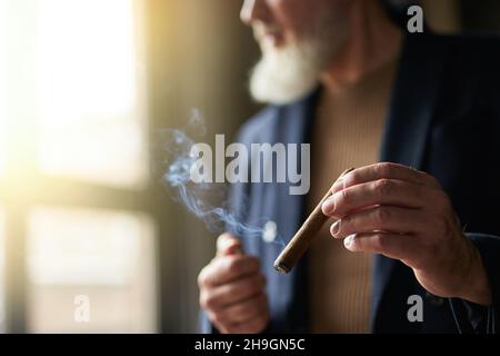 Primo piano di mani di elegante uomo maturo bearded che tiene sigaro mentre in piedi al chiuso Foto Stock