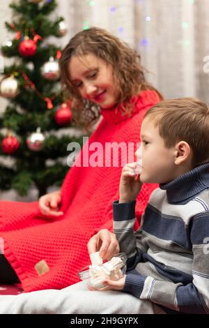 Giovane bella donna in un maglione rosso con suo figlio a casa dall'albero di Capodanno la vigilia di Capodanno. Il ragazzo sta mangiando marshmallows. Messa a fuoco selettiva. Po Foto Stock