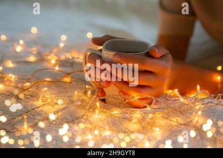 Mani donne che tengono una tazza di tè a letto. Luci di Natale accese, atmosfera accogliente casa. Foto Stock
