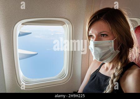 Una giovane donna in una maschera medica blu si siede nel sedile del passeggero a bordo dell'aereo e guarda fuori la finestra. Volo sicuro durante lo scoppio della co Foto Stock