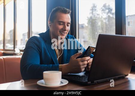 Un uomo d'affari felice uomo d'affari elegante sorridente in un attraente abito europeo funziona in un computer portatile, ascolta musica e bevande caffè seduta Foto Stock