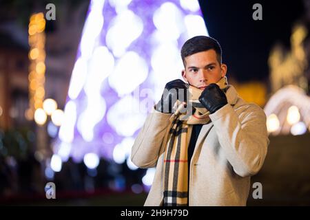 Giovane uomo che regola la sua sciarpa in inverno fuori. Albero di Natale sullo sfondo. Foto Stock
