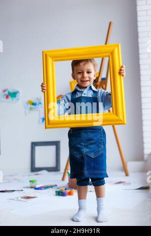 Carino, felice, ragazzo bianco in camicia blu e jeans sorridenti e guardando attraverso la cornice gialla. Piccolo bambino che si diverte in studio d'artista. Concetto di ch iniziale Foto Stock