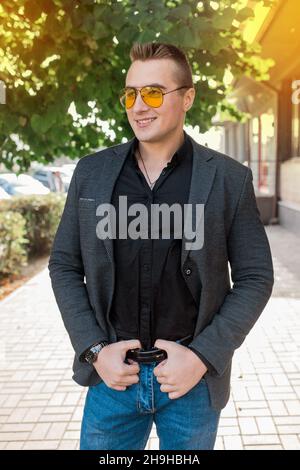 Elegante positivo sorridente giovane attraente ragazzo di aspetto europeo ritratto uomo d'affari in giacca, camicia e jeans, in occhiali da sole sulla strada outd Foto Stock