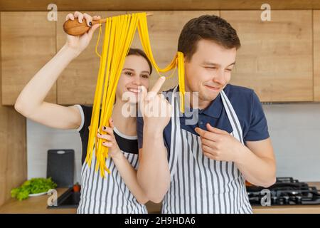 Giovane coppia in grembiuli che si diverte con tagliatelle di pasta. La famiglia cucina vegetariana italiana a casa. Concetto di stile di vita domestico, mangiare sano, felice Foto Stock