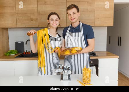Ritratto di giovane coppia in grembiuli con tagliatelle di pasta cruda. La famiglia cucina vegetariana italiana a casa. Concetto di stile di vita domestico, alimentazione sana Foto Stock