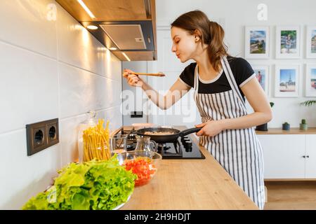 Giovane donna in grembiule cucinare cibo sano nella moderna cucina casalinga. Preparazione del pasto con padella su stufa a gas. Concetto di stile di vita domestico, ho felice Foto Stock