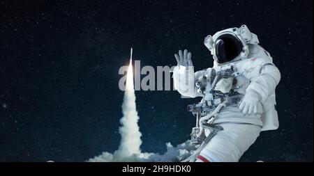 L'uomo dello spazio in un vestito e cappello dello spazio viaggia nello spazio aperto e ondeggia la sua mano contro lo sfondo di un razzo lift off. La navicella spaziale prende il via con successo Foto Stock