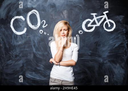 La donna bionda graziosa sceglie fra CO2 inquinamento di gas della natura e trasporto sano in bicicletta su uno sfondo di una parete del bordo del gesso. Scegliere alternativa Foto Stock